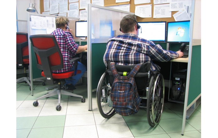 W przestrzeni biurowej, podzielonej na stanowiska oddzielone niskimi ściankami, dwie osoby pracują przy komputerach. Obie mają na głowach słuchawki z mikrofonami, co sugeruje pracę w obsłudze klienta lub centrum wsparcia. Jedna z nich siedzi na standardowym biurowym krześle, druga na wózku inwalidzkim, z przypiętym do niego plecakiem. Przed nimi ekrany monitorów, klawiatury i telefony. 