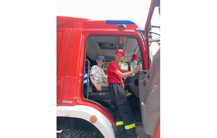 Dominik w stroju młodzieżowego strażaka zasiada za kierownicą wozu bojowego, a obok młodszy brat Bartek z uśmiechem i w stylowych okularach. Braterska drużyna gotowa na akcję – z sercem do ratowania i pasją do działania! 