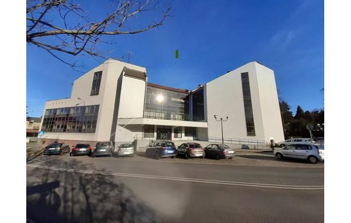 Nowoczesny, jasny budynek należący do Banku PKO BP o geometrycznej bryle góruje nad ulicą, odbijając promienie słońca w dużych przeszkleniach. Po jego bokach widoczne są wysokie, wąskie okna. Przed obiektem rozciąga się parking, gdzie zaparkowanych jest kilka samochodów. Po prawej stronie widać podjazd dla osób z niepełnosprawnościami. Nad wszystkim rozciąga się bezchmurne, błękitne niebo, a w oddali dostrzegalna jest zieleń drzew.