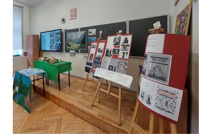 Sala lekcyjna z podwyższeniem przy tablicy. Na sztalugach ustawione są plansze z tekstami i zdjęciami, w tym reprodukcjami okładek książek i fotografiami czarno-białymi. Z boku stoi stół przykryty zielonym obrusem z makietami domów. Na ścianie widoczna jest tablica, ekran z wyświetloną prezentacją, zegar oraz godło Polski.
