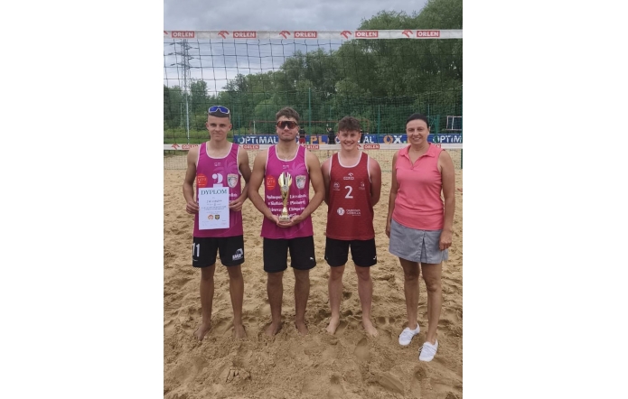 Cztery osoby stoją na boisku do siatkówki plażowej przed siatką z logo ORLEN. Dwóch chłopaków trzyma dyplom i puchar, a w tle widoczne jest piaszczyste boisko i drzewa.