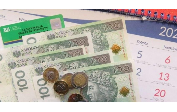 Kilka banknotów o nominale 100, ułożonych w wachlarz, oraz monety w różnych wartościach leżą na papierowej powierzchni. W tle widoczny jest fragment kalendarza z zaznaczonymi sobotami i niedzielami oraz plastikowa karta w zielonym kolorze z białym napisem, sugerująca dokument potwierdzający status emeryta lub rencisty. Spiralny grzbiet kalendarza dodaje kompozycji biurowego charakteru.