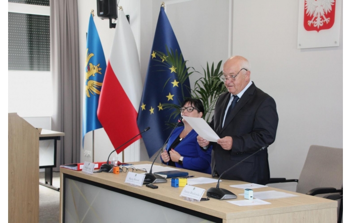 Maciej Bieniek Przewodniczący Rady w ciemnym garniturze stoi przy stole i trzyma kartkę z dokumentem. Obok niego siedzi Klaudia Kaleta Sekretarz ubrana w niebieską marynarkę. W pomieszczeniu znajdują się mikrofony, dokumenty oraz tabliczki z nazwiskami, a w tle widoczne są flagi.