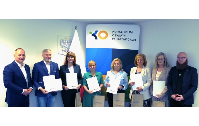 Siedem osób w eleganckich strojach stoi w rzędzie, trzymając białe teczki z logo „CFF”. W tle widoczna flaga Polski, godło oraz ścianka z napisem „Kuratorium Oświaty w Katowicach”.