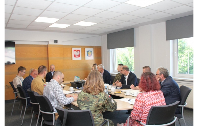 Kilkanaście osób siedzi przy okrągłym stole konferencyjnym i uczestniczy w spotkaniu. Na stole znajdują się dokumenty, notatki, filiżanki, butelki z wodą oraz sprzęt multimedialny. Wśród uczestników widoczne są osoby w stroju cywilnym oraz mundurach. Spotkanie odbywa się w jasnej sali z dużymi oknami.