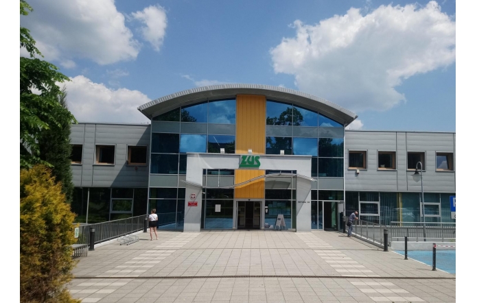 Nowoczesny budynek Zakładu Ubezpieczeń Społecznych (ZUS) z przeszkloną fasadą i charakterystycznym logo instytucji. Przed wejściem widoczna jest przestrzeń dla interesantów oraz elementy małej architektury.