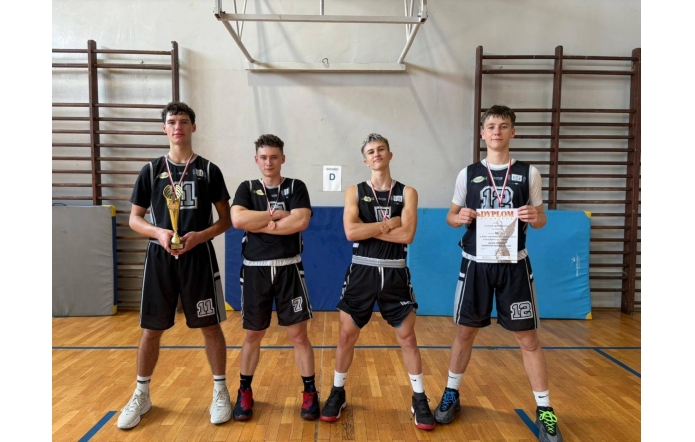 Reprezentacja szkolnej drużyny koszykarskiej po zwycięstwie – czterech zawodników w strojach sportowych prezentuje zdobyty puchar, medale oraz dyplom, stojąc dumnie na sali gimnastycznej po zakończonym turnieju.