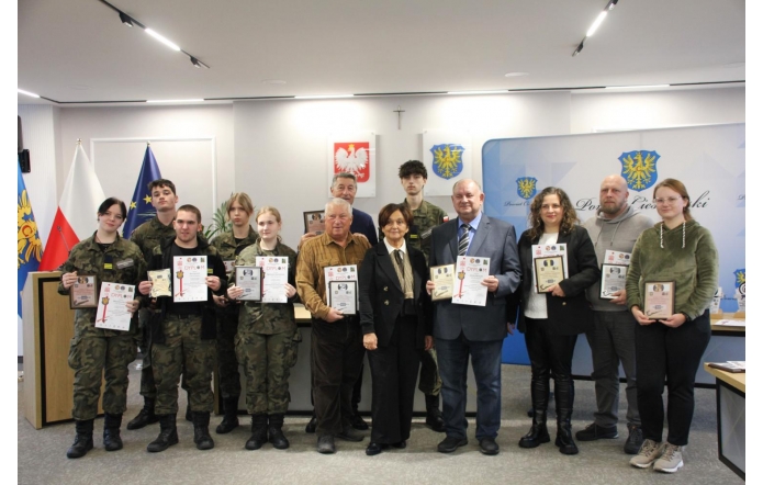 Grupa osób stoi wspólnie na sali sesyjnej, prezentując trzymane w rękach dyplomy i statuetki. W centralnej części pierwszego rzędu znajduje się Starosta Cieszyński Janina Żagan, elegancko ubrana w czarne spodnie i czarną marynarkę. Towarzyszą jej młodzi uczestnicy w mundurach oraz dorośli laureaci wyróżnień. W tle widoczne są flagi oraz godła.