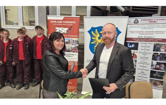 Bożena Cholewa, dyrektor Zespołu Szkół Technicznych im. płk. Gwido Langera w Cieszynie, i Marian Czyż, prezes spółki Stroirny a stavby, uśmiechnięci uściskują dłonie. W tle grupa uczniów w czerwonych uniformach i roll-upy z logami obu instytucji.