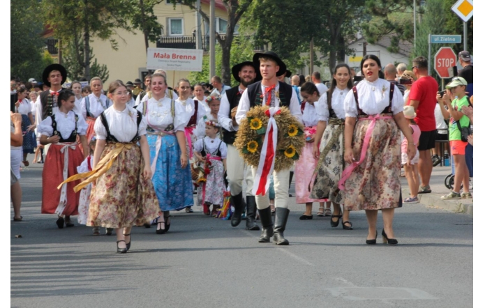 Barwny korowód uczestników Dożynek Powiatowo-Gminnych w Brennej. Grupa osób, ubrana w tradycyjne stroje ludowe, maszeruje ulicą, niosąc ozdobny wieniec dożynkowy z kwiatami słonecznika i biało-czerwoną wstążką. Kobiety mają na sobie kolorowe spódnice i bluzki z haftowanymi wzorami, a mężczyźni są ubrani w ludowe kamizelki i kapelusze. W tle widać innych uczestników korowodu oraz widownię, która obserwuje to wydarzenie. Na pierwszym planie widać również tabliczkę z napisem „Mała Brenna”, wskazującą na udział delegacji z tej miejscowości. Atmosfera zdjęcia jest radosna i pełna życia, co podkreśla tradycyjny charakter uroczystości.