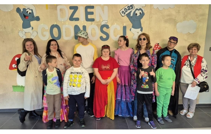 Grupa dzieci i dorosłych z Ośrodka Szkolno-Wychowawczego dla Niesłyszących i Słabo Słyszących w Raciborzu stoi przed kolorowym tłem z napisem „Dzień Godności”. Uczestnicy uśmiechają się, mają na sobie różnorodne, kolorowe stroje, a atmosfera jest radosna.