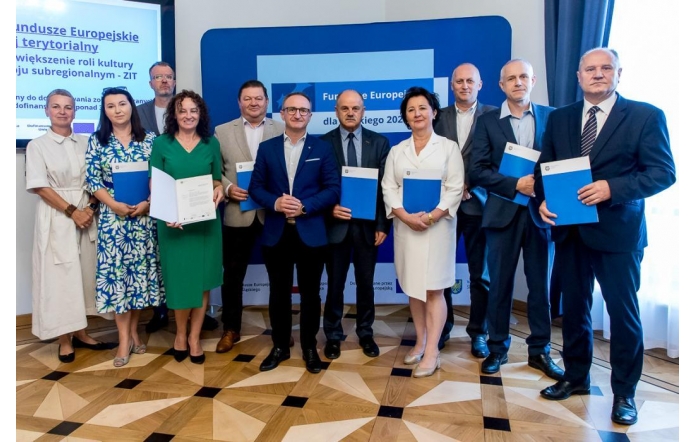 Grupa kobiet i mężczyzn w eleganckich strojach stoi na tle tablic i ekranów z logotypami Funduszy Europejskich. Część osób trzyma teczki lub dokumenty, uśmiechając się do obiektywu, co podkreśla oficjalny i uroczysty charakter spotkania.