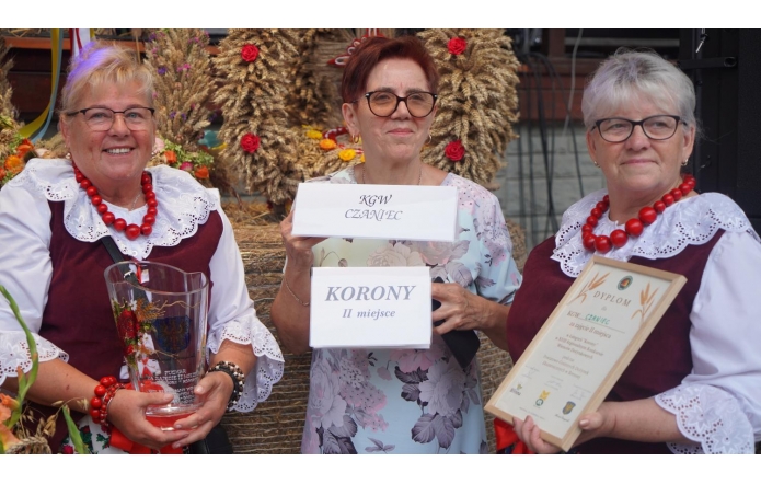 Na zdjęciu widzimy trzy kobiety, które reprezentują Koło Gospodyń Wiejskich Czaniec. Stoją one razem, dumnie prezentując swoje nagrody. Dwie z kobiet mają na sobie tradycyjne stroje ludowe, składające się z białych bluzek z koronkowymi kołnierzykami oraz ciemnoczerwonych gorsetów. Kobiety te noszą także korale w intensywnym czerwonym kolorze. Kobieta po lewej stronie trzyma w rękach szklany puchar z napisem, który wskazuje na zajęcie miejsca w konkursie. Kobieta w środku trzyma tabliczkę z napisem "KGW CZANIEC" oraz drugą tabliczkę z napisem "KORONY II miejsce", wskazującą na zajęcie drugiego miejsca w konkursie w kategorii "korony". Kobieta po prawej stronie trzyma dyplom oprawiony w ramkę, który również potwierdza zdobycie nagrody. W tle widoczne są dożynkowe wieńce wykonane z kłosów zbóż i ozdobione kolorowymi kwiatami.
