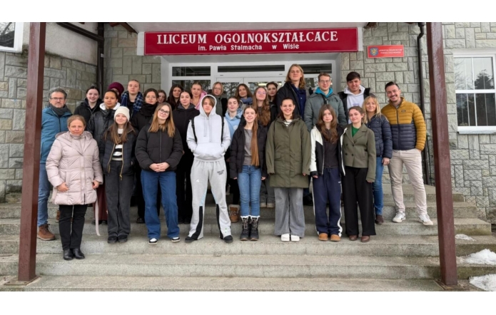 Wejście do budynku z kamienną elewacją i czerwonym szyldem „I Liceum Ogólnokształcące im. Pawła Stalmacha w Wiśle”. Na schodach stoi duża grupa młodzieży oraz kilka dorosłych osób, ubranych w kurtki i bluzy. Wszyscy pozują do wspólnego zdjęcia przed drzwiami szkoły.
