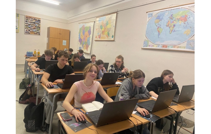 ChatGPT powiedział:  Uczniowie siedzą w sali lekcyjnej przy biurkach z laptopami, skupieni na pracy. Na ścianach wiszą mapy świata i Afryki. W tle stoją butelki z napojami. Jedna z uczennic uśmiecha się do obiektywu.