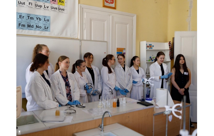 Grupa młodych kobiet w białych fartuchach laboratoryjnych stoi w sali chemicznej, przygotowana do pokazu lub prezentacji. Na ścianie widoczna tablica z układem okresowym pierwiastków i modele cząsteczek.