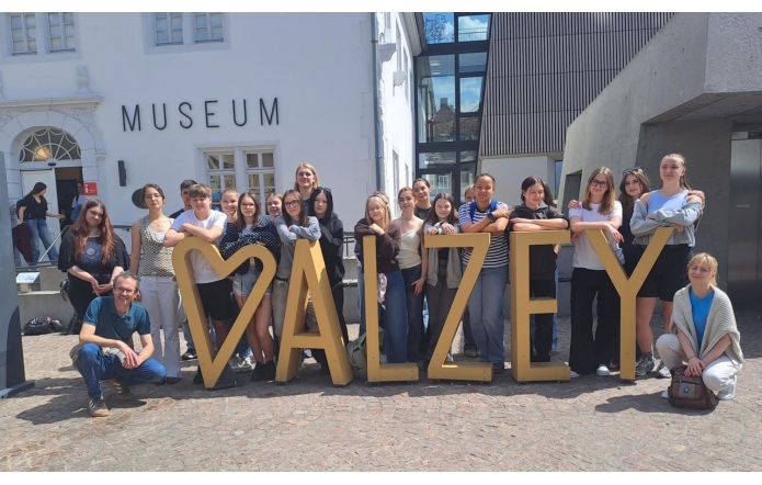 Grupa młodzieży i opiekunów pozuje przed budynkiem muzeum. Uczestnicy stoją wokół dużej przestrzennej instalacji tworzącej napis „ALZEY”, ustawionej na brukowanym placu. W tle widoczna jest jasna elewacja oraz nowoczesna zabudowa z dużymi przeszkleniami.