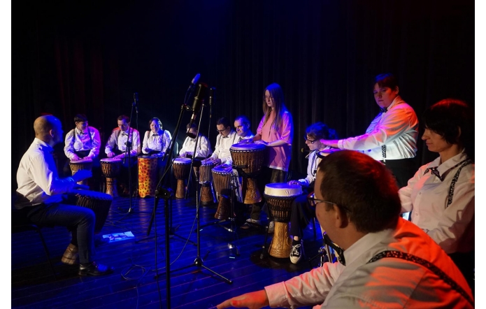 Grupa osób siedzi w półkolu na scenie, grając na bębnach, większość ubrana jest w białe koszule z muszkami, a jedna osoba w różowej bluzce stoi pośrodku i również gra, scena oświetlona jest niebieskim i czerwonym światłem