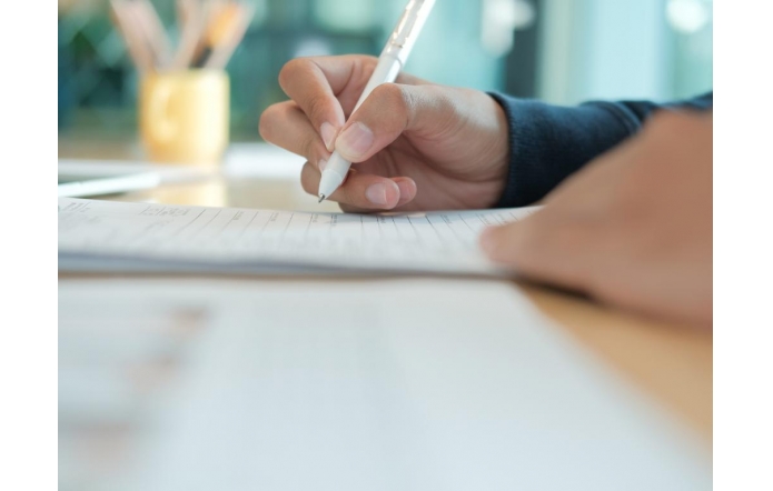 Dłoń trzymająca biały długopis pisze na kartce papieru, widoczny fragment blatu oraz rozmyte tło z przedmiotami biurowymi.