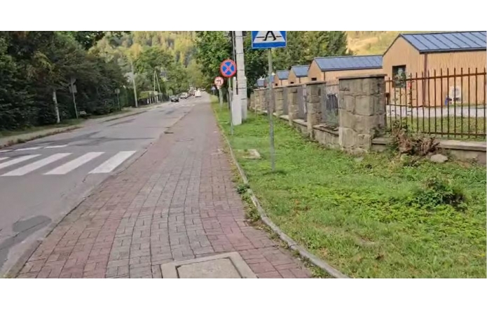 Na zdjęciu widoczny jest fragment ulicy z chodnikiem wykonanym z kostki brukowej oraz przejściem dla pieszych. Wzdłuż drogi znajdują się znaki drogowe, pas zieleni oraz ogrodzenie z kamiennymi słupkami. Po prawej stronie widoczne są budynki o jasnych elewacjach i ciemnych dachach.