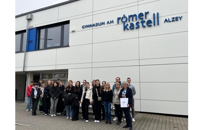 Grupa osób stoi przed budynkiem szkoły o nazwie "Gymnasium am Römerkastell Alzey". W tle widoczna jest biała elewacja z dużymi oknami. Osoby są uśmiechnięte i różnie ubrane, niektóre trzymają torby.