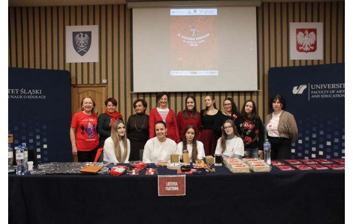 Grupa kilkunastu osób pozuje razem do zdjęcia. Z tyłu, na ekranie projektora, widoczny jest plakat 16. Cieszyńskiej Wampiriady. W pierwszym rzędzie za stołem siedzą młode osoby o, a za nimi stoi grupa kobiet w tym w odświętnych, czerwonych i zimowych stylizacjach. Po bokach widoczne są elementy scenografii z logo Uniwersytetu oraz godła.