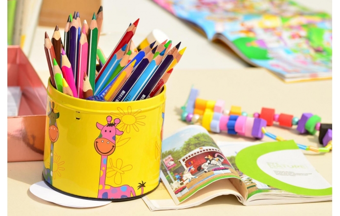 Metalowy kubek z kolorowymi kredkami, obok otwarta książka i kolorowe koraliki na stole.