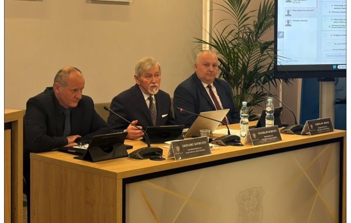 Po lewej stronie siedzi Grzegorz Konieczny Wiceprzewodniczący Rady ubrany w czarny garnitur, obok niego Stanisław Kubicius Przewodniczący Rady w granatowym garniturze, a następnie Jarosław Bodak Wiceprzewodniczący Rady w niebieskim garniturze. Przed nimi stoją tabliczki z imionami i nazwiskami, tablety oraz laptop, a także butelki z wodą i szklanki. Po prawej stronie widoczny jest ekran z wyświetlonym porządkiem obrad, w tle znajduje się roślina doniczkowa.