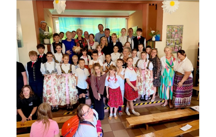 Do wspólnego zdjęcia pozują dzieci, młodzież oraz osoby dorosłe. Część uczestników ubrana jest w stroje regionalne. Grupa ustawiona jest na scenie na tle dekoracji przedstawiającej krajobraz.