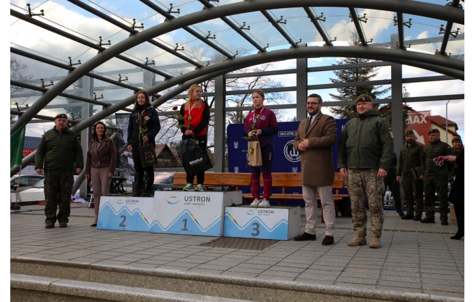 Trzy kobiety stoją na podium zwyciężczyń zawodów sportowych w Ustroniu; otrzymują róże i upominki. Obok nich stoją uśmiechnięci przedstawiciele wojska i władz lokalnych w otwartej, zadaszonej przestrzeni miejskiej.