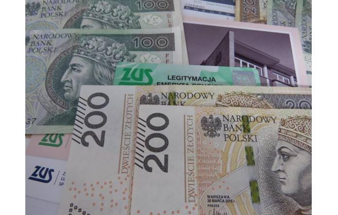 Rozłożone są banknoty o nominałach 100 i 200 złotych, a pomiędzy nimi leżą dokumenty i karty z logo ZUS, w tym zielona legitymacja emeryta-rencisty. Kolory tworzą kontrast zieleni, bieli i beżu, a w tle widać fragment fotografii budynku.