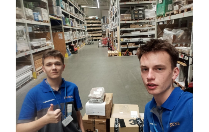 Dwóch młodych mężczyzn w niebieskich koszulkach z identyfikatorami znajduje się w magazynie pełnym regałów z towarem. Jeden z nich pokazuje kciuk w górę, a obok nich stoją kartony przygotowane do wysyłki.
