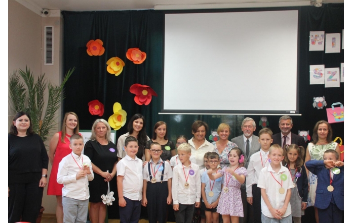 Na zdjęciu widać grupę dzieci i dorosłych pozujących razem w sali ozdobionej kolorowymi papierowymi kwiatami. Dzieci stoją z przodu, część ma na szyi medale i odznaki, a dorośli –  nauczyciele i goście – stoją za nimi, uśmiechając się do zdjęcia.