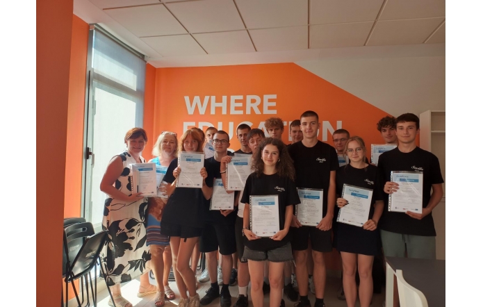 Grupa młodych osób wraz z nauczycielkami pozuje do zdjęcia w jasnym pomieszczeniu z pomarańczową ścianą z napisem. Wszyscy trzymają w rękach certyfikaty, uśmiechając się do kamery.