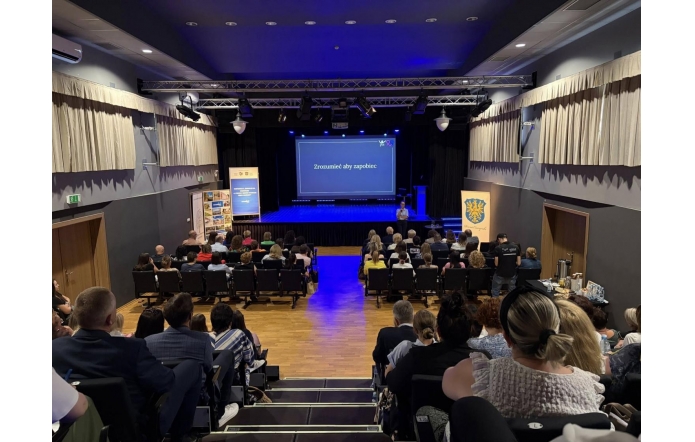 Uczestnicy siedzą na widowni sali konferencyjnej skierowanej w stronę sceny. Na scenie wyświetlana jest prezentacja, a przed ekranem znajduje się osoba prowadząca wystąpienie. Po bokach sceny ustawiono banery, a sala jest wypełniona publicznością.
