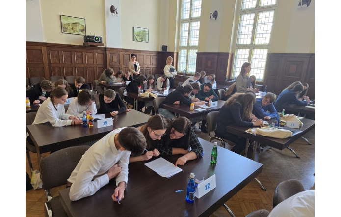 Zdjęcie przedstawia grupę młodych ludzi w eleganckich strojach siedzących przy stołach i pracujących nad zadaniami w parach lub małych zespołach. W tle znajdują się osoby nadzorujące przebieg wydarzenia. Sala jest przestronna, z dużymi oknami i drewnianymi elementami wystroju. Na stołach widoczne są numerki oznaczające drużyny, butelki z wodą i inne materiały pomocnicze. Atmosfera skupienia i zaangażowania sugeruje, że uczestnicy biorą udział w konkursie lub olimpiadzie naukowej.