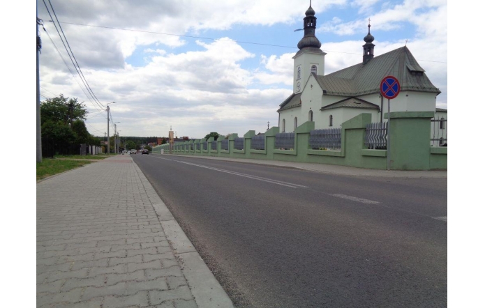Droga asfaltowa biegnie obok kościoła z dwiema wieżami, otoczonego zielonym murem. W tle widać domy i drzewa pod pochmurnym niebem. Po lewej stronie drogi znajduje się chodnik z betonowych płyt.