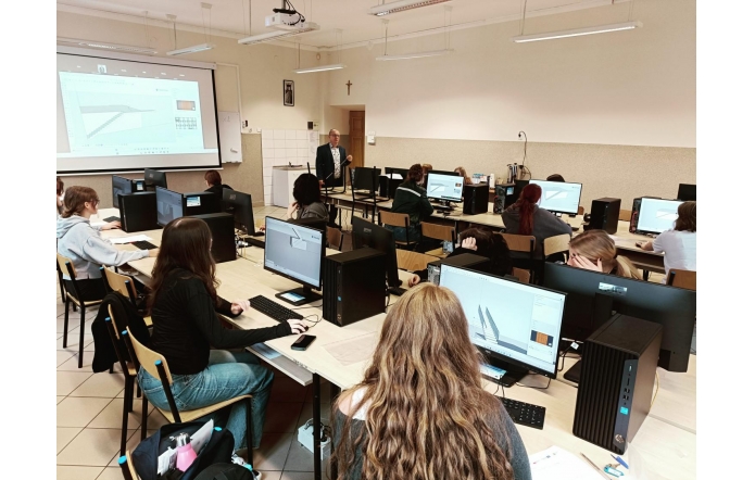 Sala komputerowa, w której uczniowie siedzą przy biurkach i pracują na komputerach. Na monitorach otwarty jest program do projektowania 3D, a na ścianie wyświetlony jest obraz z projektora z tym samym programem. Przy tablicy stoi nauczyciel.