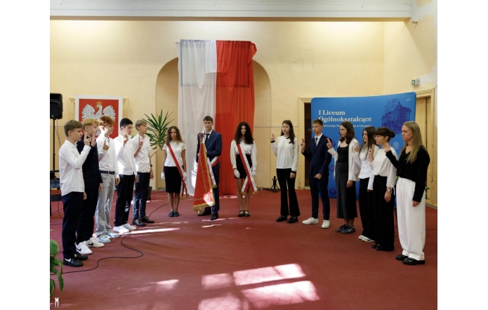 Grupa uczniów w strojach galowych stoi w sali z polskimi flagami, przygotowana do występu lub apelu.
