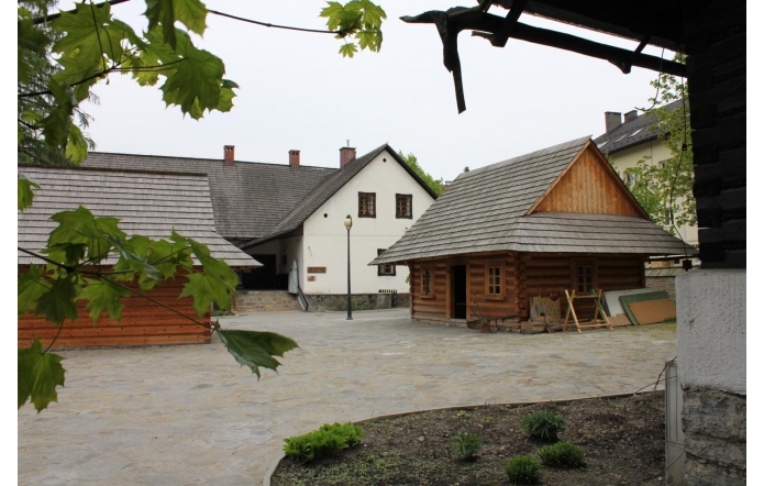 Dziedziniec z kamienną nawierzchnią otoczony drewnianymi obiektami z gontowymi dachami oraz jasnym budynkiem z kamienną podmurówką. Przy ścianach ustawione są drewniane elementy i tablice, w przestrzeni znajduje się latarnia, a w kadrze widoczne są także liście drzew.