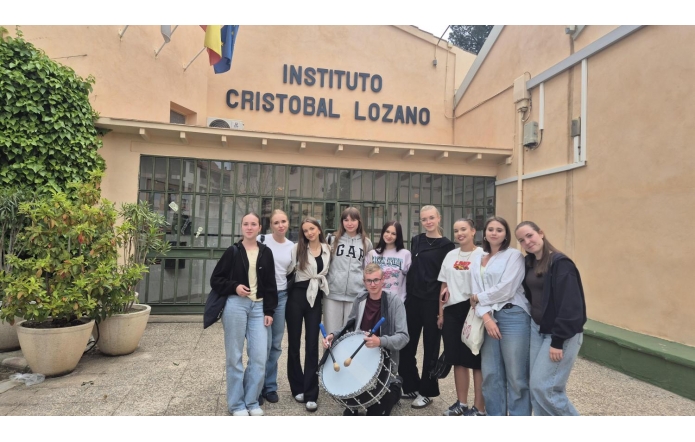 Grupa młodych osób stoi przed budynkiem z napisem "Instituto Cristobal Lozano". W centrum grupy znajduje się osoba z bębnem. W tle widać rośliny w donicach i flagi na dachu. Uczestnicy są uśmiechnięci i ubrani na sportowo.