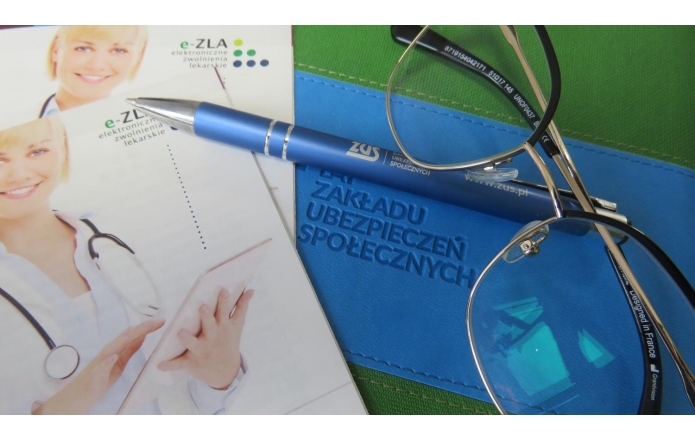 Ulotka z lekarką i napisem „e-ZLA – elektroniczne zwolnienia lekarskie”, obok niebieskie etui i długopis z logo ZUS, na których leżą okulary w metalowych oprawkach.