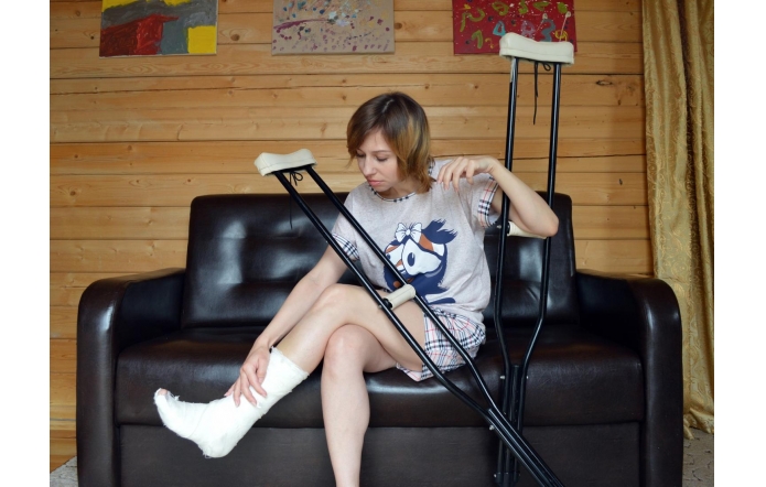 Kobieta w piżamie siedzi na ciemnobrązowej skórzanej kanapie, opierając się o nią i trzymając kule ortopedyczne. Ma nogę w gipsie, którą delikatnie dotyka. Tło stanowi drewniana ściana z trzema kolorowymi obrazami, a po prawej stronie widać beżową zasłonę.