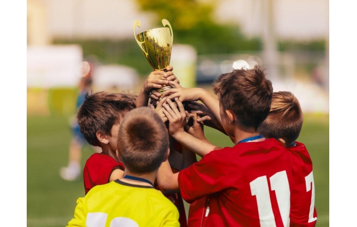 Grupa małych chłopców na boisku unosi razem złoty puchar. Czerwone koszulki piłkarskie, numery na plecach, medale na szyjach; jeden w żółtej bluzie. W tle rozmyta murawa i jasny dzień, euforia po wygranej. Dłonie na pucharze, miny skupione. W górze!