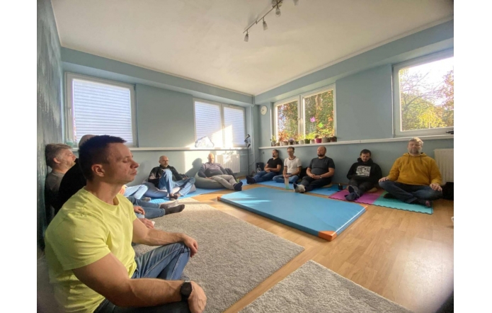 Grupa mężczyzn siedzi w kręgu na matach i dywanach w niebieskiej sali z dużymi oknami. Przez okna wpada światło słoneczne, oświetlając wnętrze. Na parapetach stoją doniczki z roślinami, a na podłodze leżą kolorowe maty i miękkie dywany.