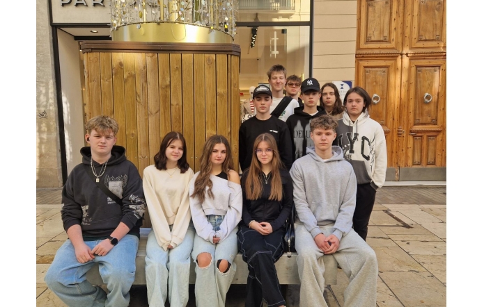 Dziewięć młodych osób ustawionych w dwóch rzędach – część siedzi na ławce, pozostali stoją za nimi. Ubrani w codzienne stroje w stonowanych kolorach. W tle drewniana zabudowa, duże drzwi z ozdobnymi uchwytami oraz przeszklone wejście do budynku.