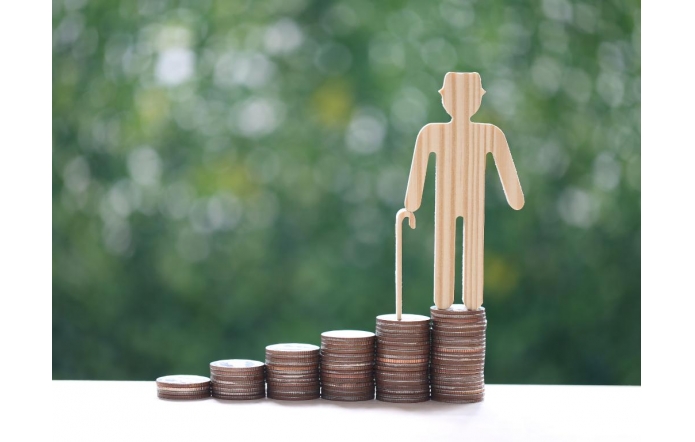 Drewniana figurka starszej osoby z laską stoi na najwyższym stosie monet, obok kilku niższych stosów ułożonych w rosnącym rzędzie. Tło jest rozmyte, zielone, przypominające liście drzew.