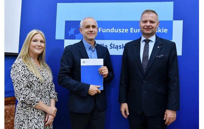 Trzy osoby stoją przed niebieską ścianką z napisem „Fundusze Europejskie dla Śląskiego”. Mężczyzna w środku trzyma w ręku niebieską teczkę z logo. Wszyscy ubrani są w stroje formalne – dwaj mężczyźni w garniturach, kobieta w jasnej wzorzystej sukience – i pozują do zdjęcia.