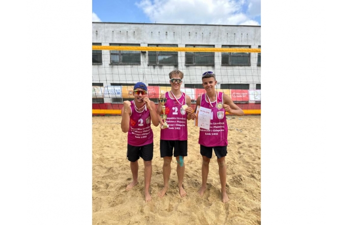 Trzech młodych siatkarzy plażowych w różowych koszulkach świętuje zwycięstwo – trzymają puchar, medale i dyplom, stojąc boso na piasku przed siatką i budynkiem sportowym.