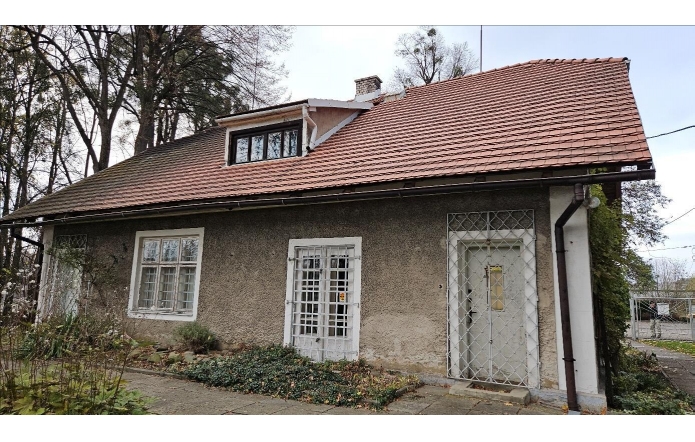 Domek Ogrodnika – Muzeum Zofii Kossak-Szatkowskiej w Górkach Wielkich od strony dawnego parku dworskiego, niewielki historyczny budynek z jasną elewacją i spadzistym, ceglanym dachem, otoczony zielenią.